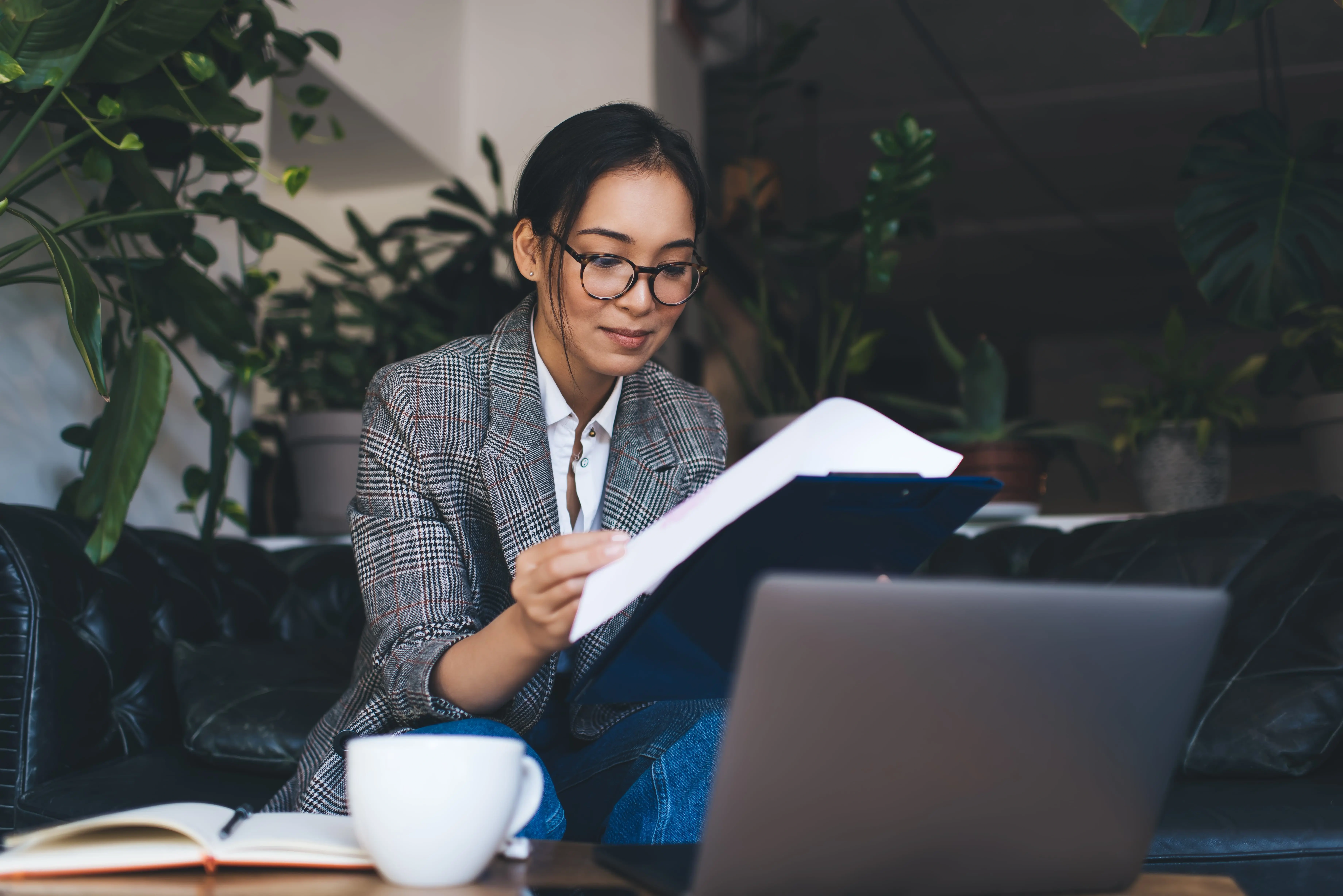 Young Asian women in glasses reading a folder at a sofa with a laptop open in front of her at the coffee table