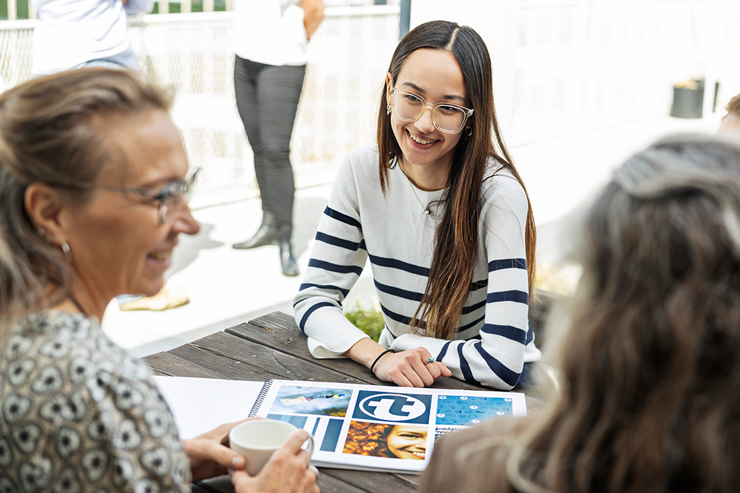 Tre smilende damer sidder ved et bord udenfor og holder et møde med printet materiale på bordet.
